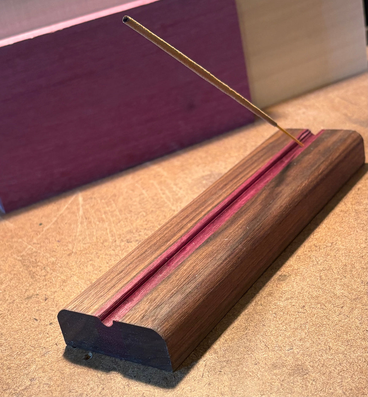 A wooden incense holder with a pink-lined slot holding a single burning incense stick, photographed on a tan textured surface with a purple block in the background.