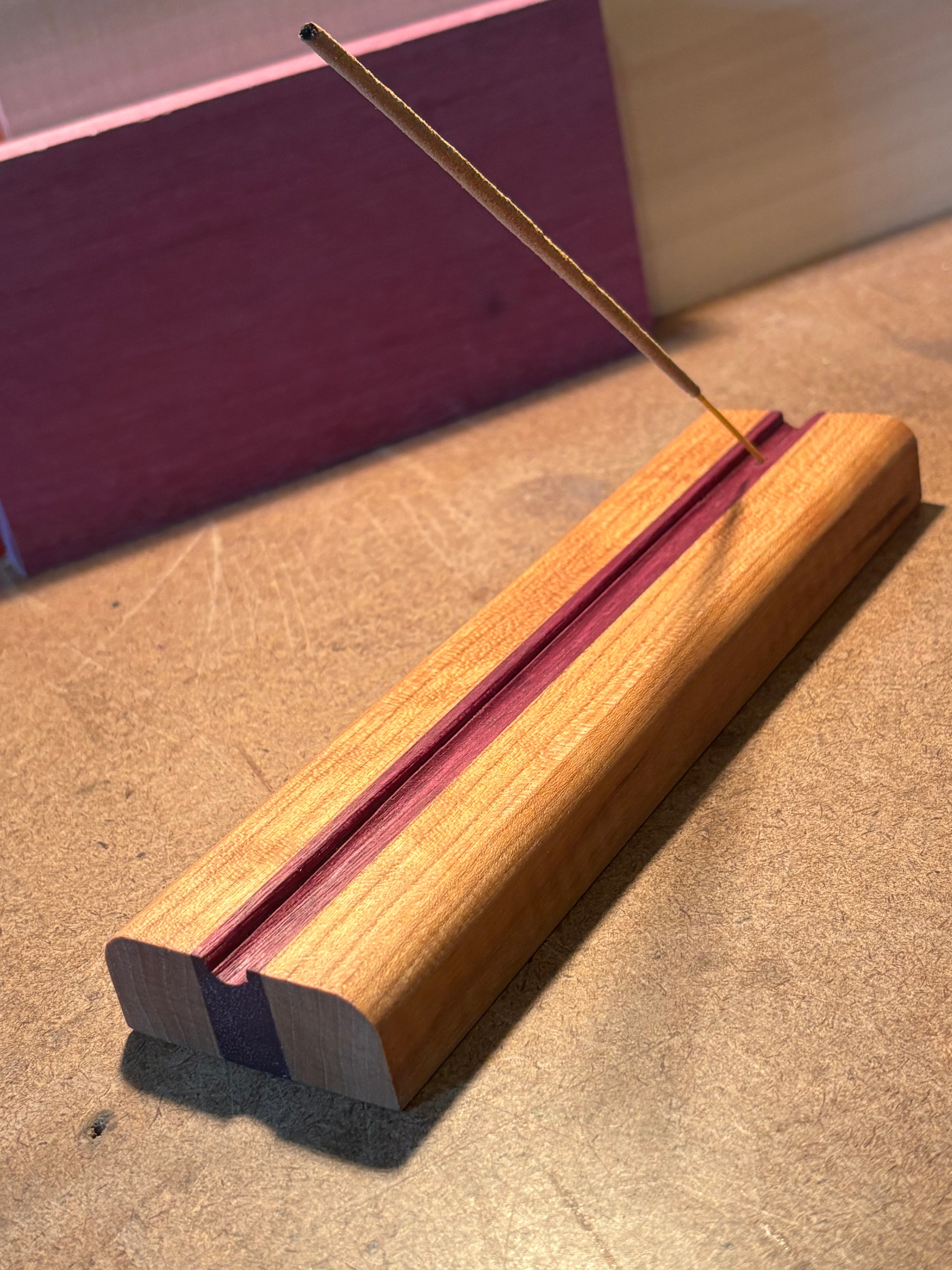 A wooden incense holder with a reddish-pink center stripe and a single incense stick resting diagonally across it, photographed on a tan textured surface.
