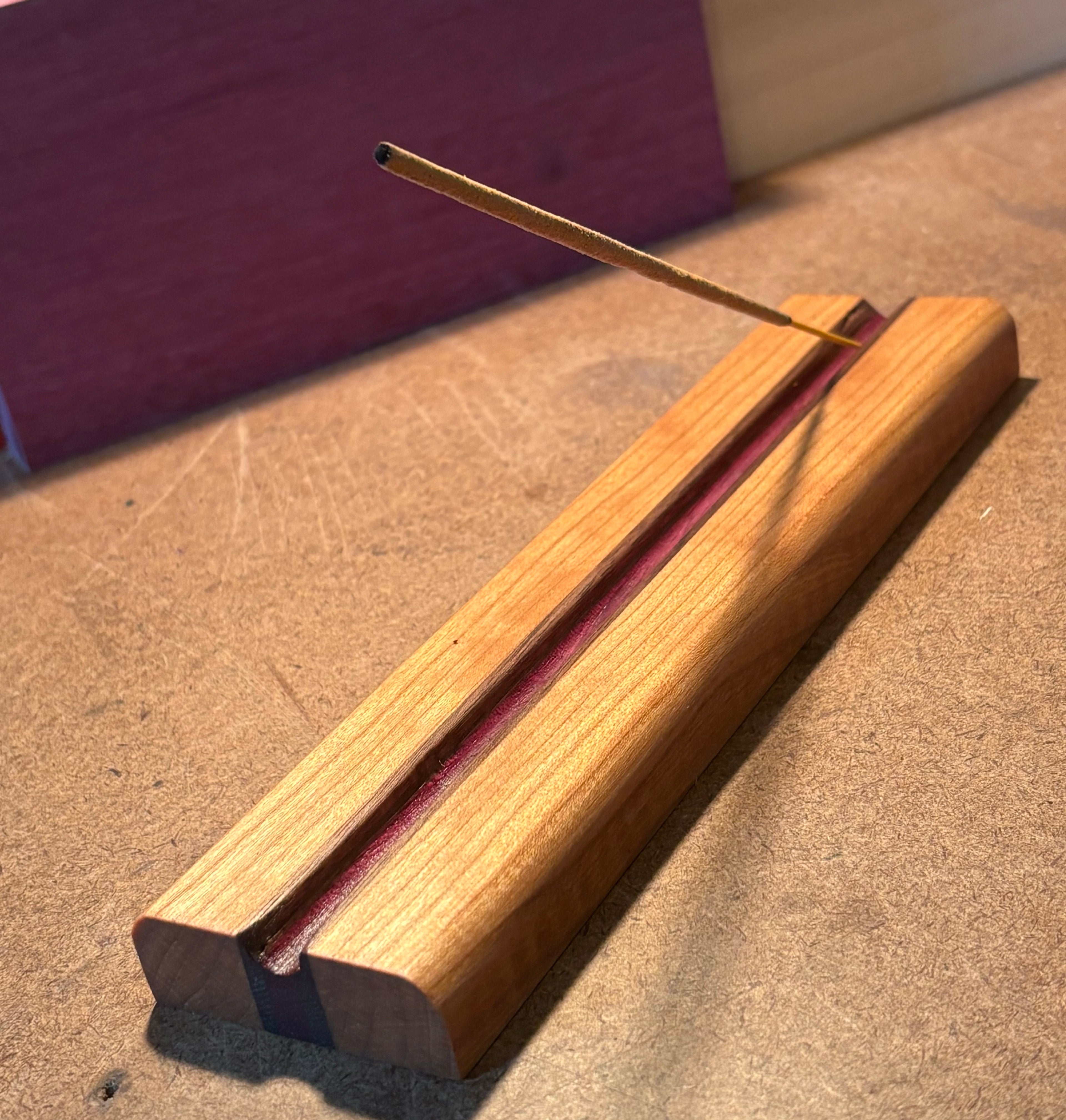 A wooden incense holder with a single incense stick resting diagonally across the top slot, photographed on a tan tabletop with a blurred purple background.