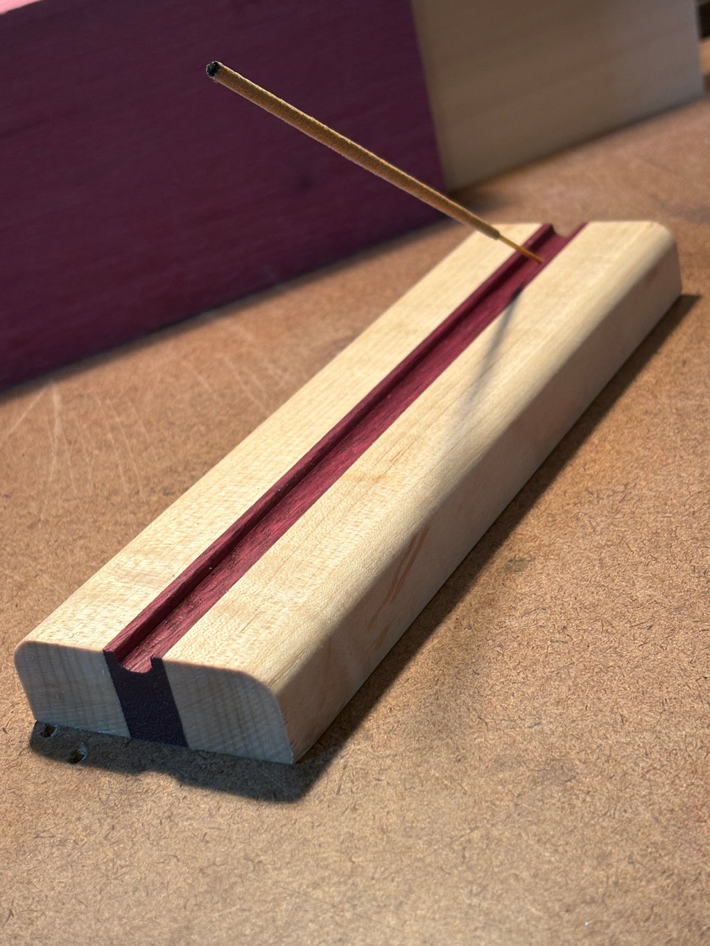 A close-up of a wooden incense holder with a light natural wood outer layer, a reddish-brown inner layer, and a dark purple stripe, shown with a thin incense stick extending from the left side against a textured brown background.