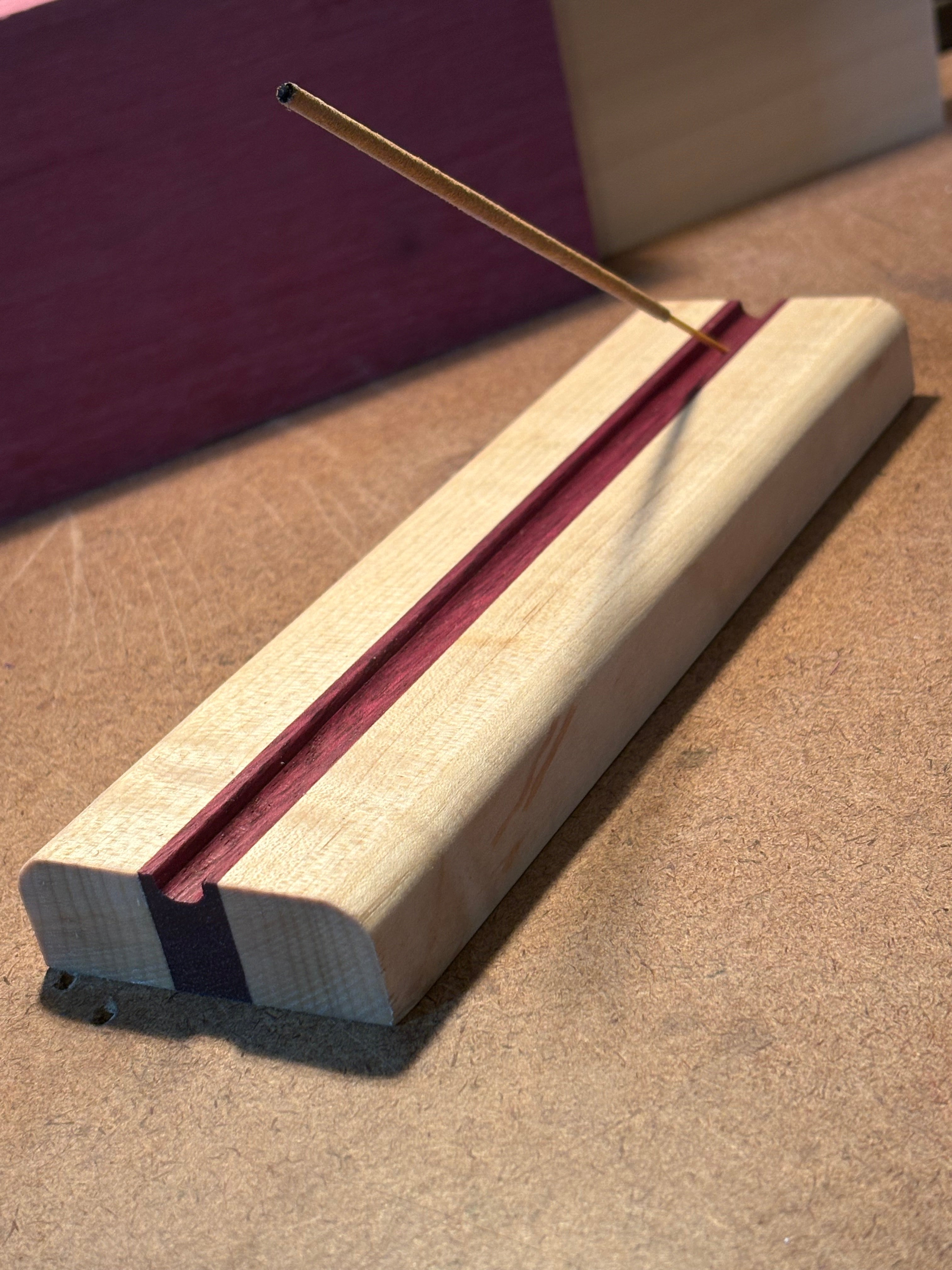A close-up of a wooden incense holder with a light natural wood outer layer, a reddish-brown inner layer, and a dark purple stripe, shown with a thin incense stick extending from the left side against a textured brown background.