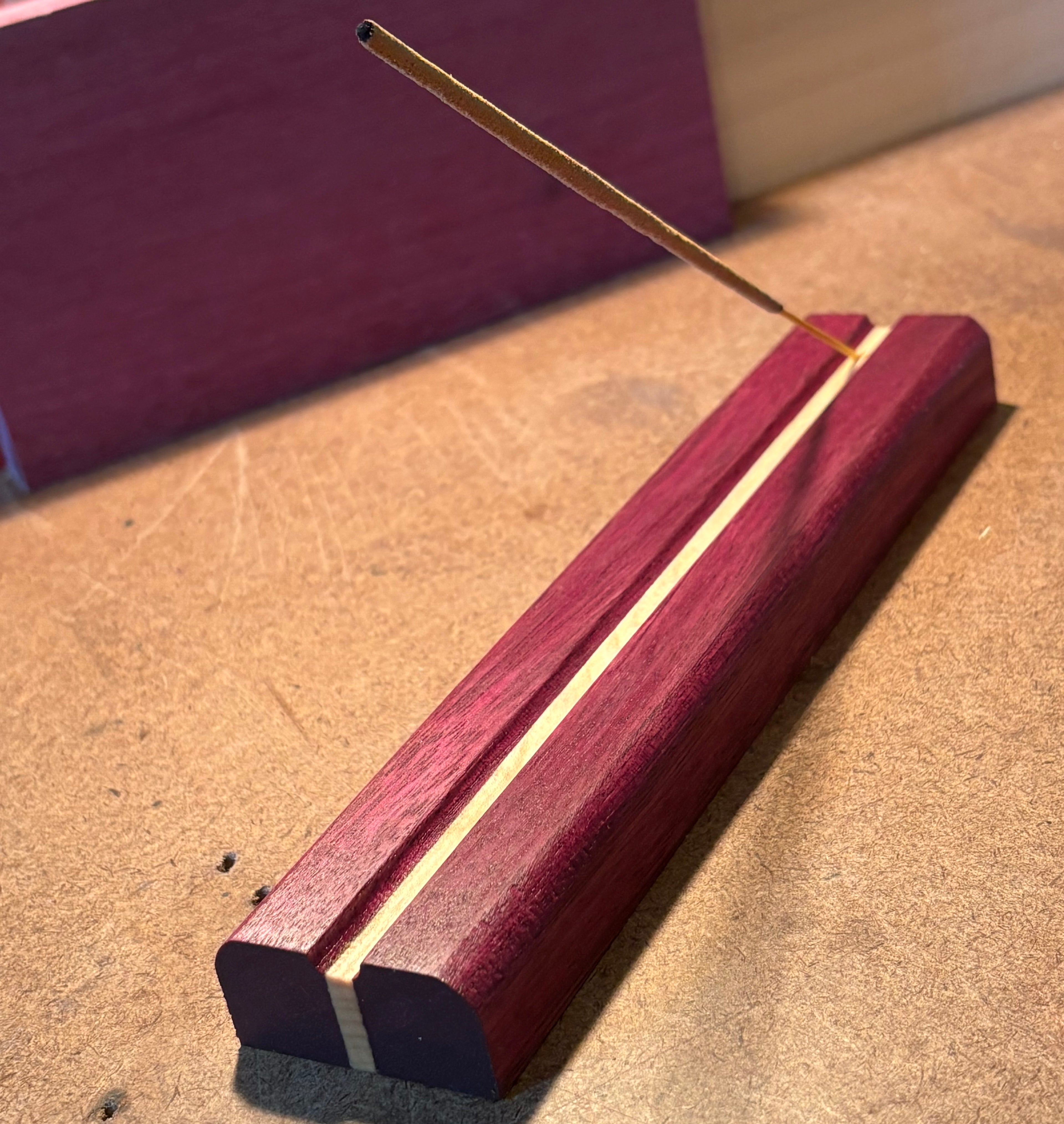 A close-up of a wooden incense holder with a single incense stick resting diagonally across the top slot, set on a tan textured surface.