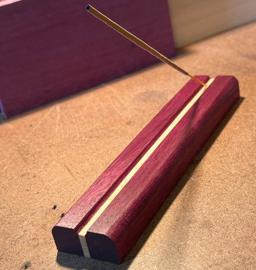 A close-up of a wooden incense holder with a single incense stick resting diagonally across the top slot, set on a tan textured surface.